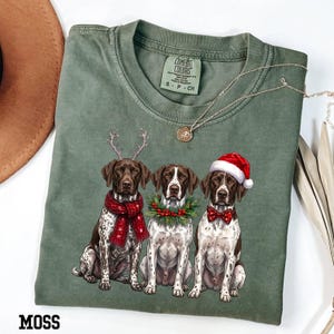 Op de afbeelding: Een mosgroen t-shirt met ronde hals en een afbeelding van drie Duitse staande honden. De linkerhond draagt een rode sjaal en rendiergeweien, de middelste hond heeft een hulstkrans en de rechterhond draagt een rode kerstmuts en strik.