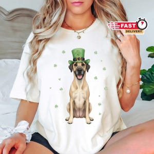Könnte beinhalten: Cremefarbenes T-Shirt mit einem hellbraunen Hund, der einen grünen St. Patrick's Day Hut trägt. Der Hund sitzt mit herausgestreckter Zunge, umgeben von grünen Kleeblatt-Grafiken. Das Shirt hat einen Rundhalsausschnitt und kurze Ärmel.