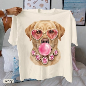 May include: An off-white t-shirt featuring a graphic of a golden retriever wearing pink heart-shaped sunglasses and a pink bubblegum bubble in its mouth. The dog is adorned with a gold-toned necklace featuring large, faceted pink oval gemstones.
