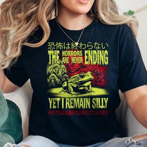 May include: Black t-shirt featuring a green and red graphic of a frog and the text "The horrors are never ending, yet I remain silly". The text is in Japanese and English.