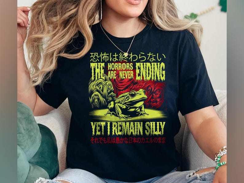 May include: Black t-shirt featuring a green and red graphic of a frog and the text "The horrors are never ending, yet I remain silly". The text is in Japanese and English.