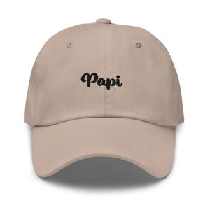 May include: A beige baseball cap featuring the word Papi embroidered in black cursive script across the front centre. The hat has a structured crown, a curved brim, and a button at the top, with visible stitching details along the panels and brim.