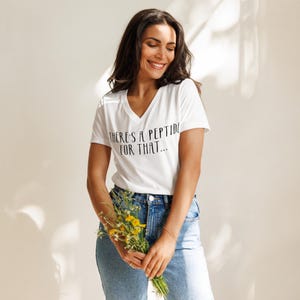 Peut inclure: Un t-shirt blanc à col en V avec le texte "THERE'S A PEPTIDE FOR THAT..." en noir. La personne porte un jean bleu et tient un bouquet de fleurs jaunes. La personne sourit.