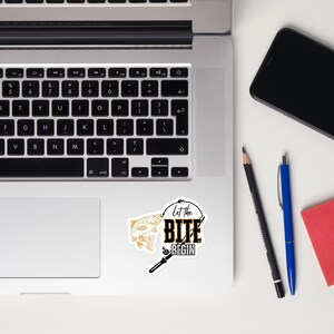 Può includere: Un adesivo a tema pesca. Presenta un pesce dorato che salta fuori dall'acqua, una canna da pesca e il testo "Let the BITE BEGIN" in nero e oro. L'adesivo è su una superficie bianca.