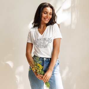 Peut inclure: Un t-shirt blanc à col en V avec le texte "PEPTIDE LEVEL PRECISION" en noir. Le t-shirt est porté avec un jean bleu. La personne tient un bouquet de fleurs jaunes.
