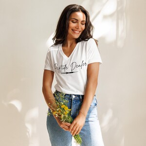Peut inclure: T-shirt blanc à col en V avec l'inscription "Peptide Dealer" en écriture noire et un dessin de seringue. La personne porte un jean bleu et tient un bouquet de fleurs jaunes. Le t-shirt est de style décontracté.