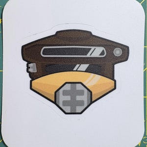 May include: A sticker with a brown and yellow helmet design. The helmet has a rectangular visor and a geometric shape on the lower section. The sticker is on a white background.