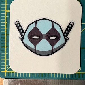 May include: A sticker featuring a stylized mask design in light blue, gray, and black, with two katana swords on either side. The mask has diamond-shaped eye openings and a central vertical stripe. The sticker is on a white, square-cornered background.
