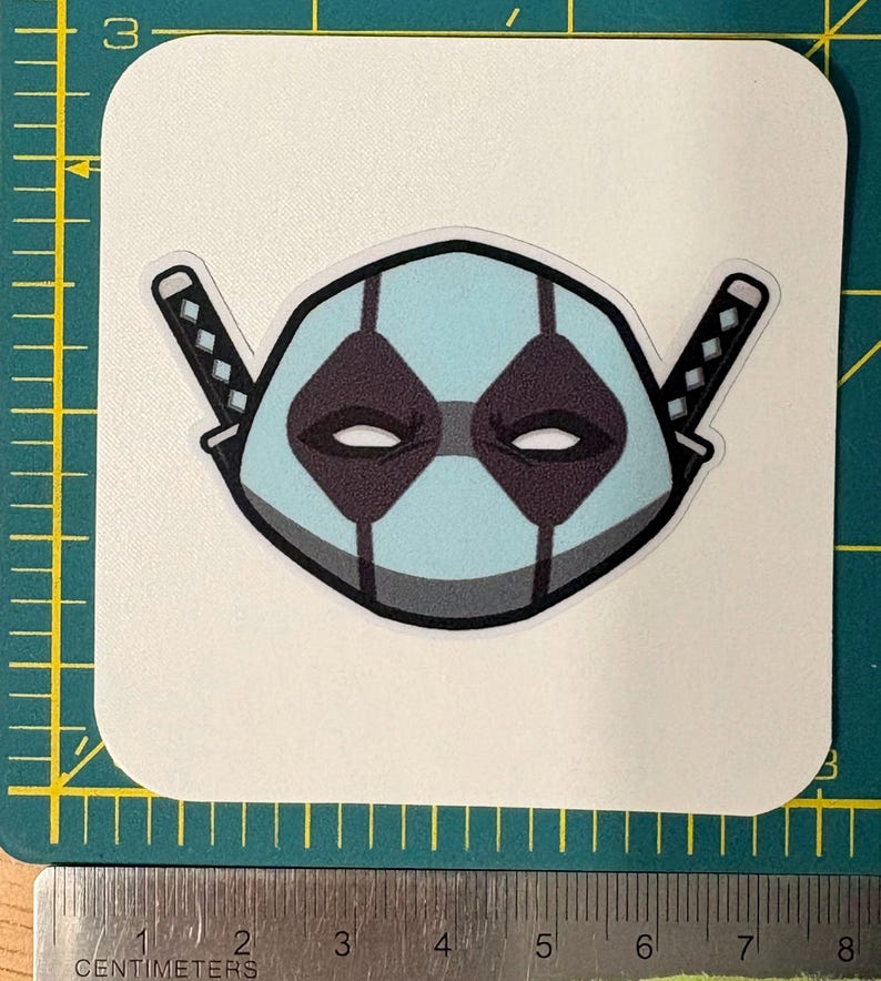 May include: A sticker featuring a stylized mask design in light blue, gray, and black, with two katana swords on either side. The mask has diamond-shaped eye openings and a central vertical stripe. The sticker is on a white, square-cornered background.