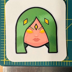 May include: A sticker featuring a stylized illustration of a female face with green hair and yellow eyes. The face has a light skin tone and is framed by green hair with yellow diamond accents. The sticker is on a green cutting mat with a ruler.