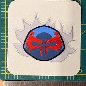 Może przedstawiać: Naklejka z wystylizowanym emblematem z niebieskim tłem i czerwonym wzorem. Emblemat otoczony jest szarym efektem rozbłysku na białym tle. Naklejka ma około 6,35 cm szerokości.