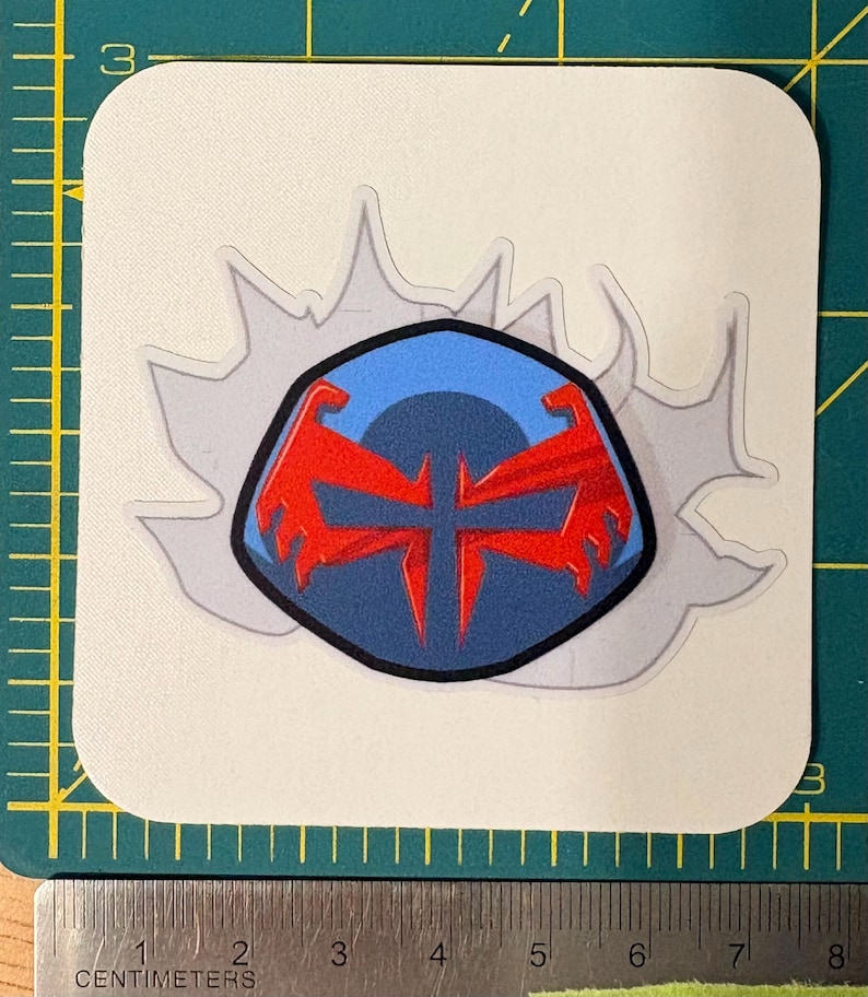 Może przedstawiać: Naklejka z wystylizowanym emblematem z niebieskim tłem i czerwonym wzorem. Emblemat otoczony jest szarym efektem rozbłysku na białym tle. Naklejka ma około 6,35 cm szerokości.