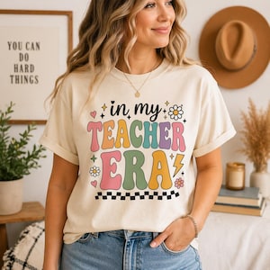 Könnte beinhalten: Cremefarbenes T-Shirt mit dem Text "in my TEACHER ERA" in Retro-Schrift. Das Design umfasst Herzen, Blumen und einen Blitz. Das Shirt wird von einer Person in Jeans getragen.