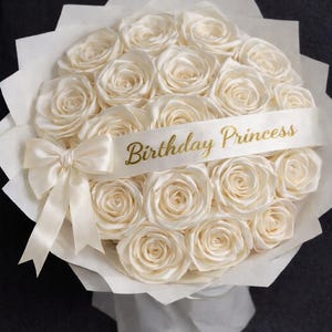 May include: A bouquet of cream-coloured roses, wrapped in white paper, and tied with a matching ribbon. A banner across the flowers reads "Birthday Princess" in gold lettering. The arrangement is set against a dark background.