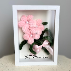 Puede incluir: Una caja de sombra enmarcada en blanco con un ramo de flores rosas en su interior. Las flores están hechas de material rosa peludo y tienen detalles de perlas blancas. El texto "Best Mom Ever" está escrito en cursiva negra en la parte inferior de la caja.