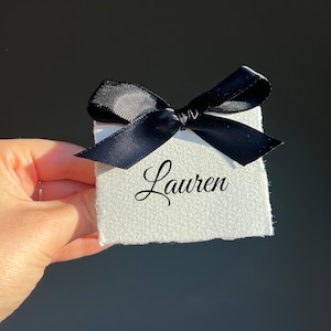 Op de afbeelding: Een kleine, witte, getextureerde geschenkdoos, vastgebonden met een zwarte satijnen strik. De naam "Lauren" is in elegant schrift op de voorkant van de doos geschreven. De doos wordt in een hand tegen een donkere achtergrond gehouden.