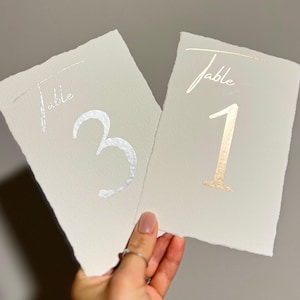Peut inclure: Deux cartons de numéros de table blanc cassé. Le mot "Table" est imprimé en écriture manuscrite en haut de chaque carton. L'un porte le numéro "3" en feuille d'argent, l'autre le numéro "1" en feuille d'or.