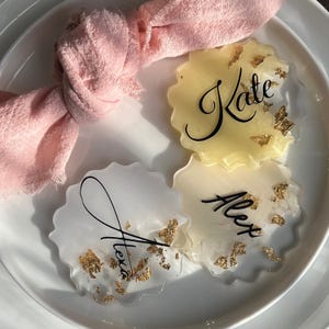 May include: Three resin coasters with scalloped edges, featuring gold leaf accents and personalized names in black script. One coaster is yellow, and two are white. A pink cloth napkin is in the background.
