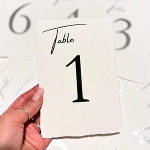 Könnte beinhalten: Cremefarbene Tischkarten mit dem Wort „Table“ in Schreibschrift über einer großen schwarzen Zahl 1. Die Karte hat einen strukturierten, gezackten Rand. Weitere Tischkarten sind im Hintergrund zu sehen.