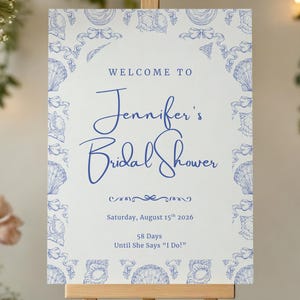 May include: A white sign on a wooden easel with blue text that reads "WELCOME TO Jennifer's Bridal Shower". The sign is decorated with a blue floral border and includes the date, August 15th, 2026. Floral arrangements and candles are in the background.