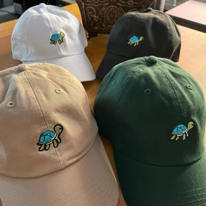 Pode incluir: Quatro bonés de beisebol com um pequeno desenho de tartaruga bordado. Os bonés são de cor branca, preta, bege e verde.