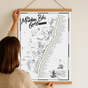 May include: A monochrome map titled "The Mountain Bike Quest NSW & ACT" is displayed in a wooden frame. The map features illustrations of mountain bikers and a detailed route. The map is being held up by a person.