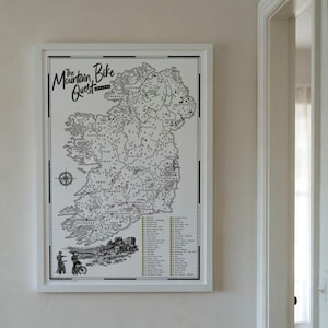 May include: A framed white wall art print titled The Mountain Bike Quest featuring a detailed black and white map of Ireland. The map includes a compass rose, illustrations of two mountain bikers, and a numbered list of locations in yellow.
