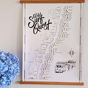 May include: A white wall hanging poster titled NSW Surf Quest featuring a black ink map of the New South Wales coastline, a list of surf locations with gold circular markers, a compass rose, and a drawing of a van and surfers, held by wood rails.
