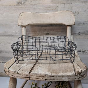 May include: A miniature wire sofa with rounded armrests sits on a distressed, white-painted wooden chair. The sofa is constructed from dark gray wire, creating a cage-like appearance. The chair has a rustic, aged aesthetic.