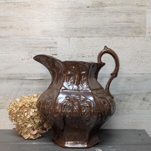 May include: A brown ceramic pitcher with a handle and a spout. The pitcher has a detailed, embossed floral design and sits on a dark gray surface. A dried flower arrangement is next to the pitcher.