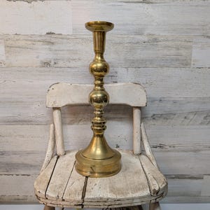 May include: A tall, brass candlestick holder with a round base, a central column with two spherical accents, and a top cup for holding a candle. The candlestick is displayed on a distressed white wooden chair against a white and gray wood-paneled wall.