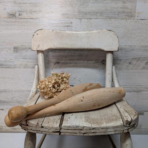 May include: Two wooden antique exercise clubs rest on a distressed white chair. The clubs are light brown with rounded ends. A bundle of dried flowers sits behind the clubs. The chair has a weathered, vintage appearance.