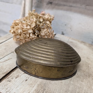 May include: A vintage brass trinket box with a lid featuring concentric circular ridges. The box has a rounded, clam-shell-like shape and a slightly aged patina. A dried flower arrangement is in the background, adding a touch of rustic charm.
