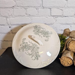 May include: A white ceramic serving dish lid with a floral and scroll design in gray. The lid has a small handle and a scalloped edge. The dish is on a black surface with a white brick background.