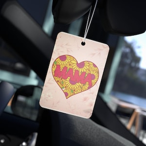 May include: Pink car air freshener with a heart-shaped design featuring yellow sunflowers and the word "MAMA" in red letters.