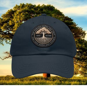 May include: A navy blue baseball cap featuring a circular patch with a tree design. The patch includes the phrases "Your branches define your reach", "Your roots define your strength", "DIG DEEP", and "HOLD STRONG". The background shows a large tree and a blue sky.