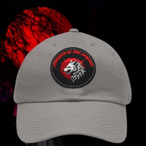 Könnte beinhalten: Graue Baseballkappe mit einem runden Aufnäher mit einem weißen Wolfskopf in einem roten Kreis, mit dem Text "STRENGTH IN THE SHADOWS". Der Hintergrund zeigt einen roten Mond und eine dunkle Silhouette.