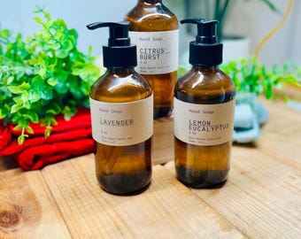 May include: Three amber glass bottles of hand soap with black pump dispensers. Each bottle features a tan label with black text. The labels read Citrus Burst 8 oz, Lavender 8 oz, and Lemon Eucalyptus 8 oz, all by Bare Naked Salts and Soap, LLC.