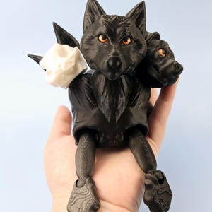 May include: A black three-headed wolf figurine with orange eyes and a white skull head. The figurine is held in a hand against a light blue background. The wolf has articulated legs and a textured surface.