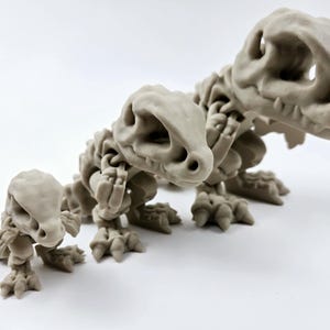 Könnte beinhalten: Drei graue Skelett-Dinosaurier-Figuren in verschiedenen Größen. Der größte Dinosaurier steht auf allen Vieren mit erhobenem Kopf. Der mittlere Dinosaurier steht auf allen Vieren mit gesenktem Kopf. Der kleinste Dinosaurier sitzt auf seinen Hinterbeinen mit erhobenem Kopf.
