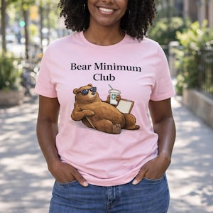 May include: A light pink t-shirt with the text "Bear Minimum Club" and a cartoon bear wearing sunglasses and holding a coffee cup. The person is wearing blue jeans.