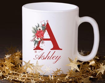 Op de afbeelding: Witte keramische mok met een rode letter "A" en de naam "Ashley" in rood schrift. De letter is versierd met een bloemstuk van rode poinsettia's, groene bladeren en sneeuwvlokken. De mok is omgeven door gouden stervormige decoraties.