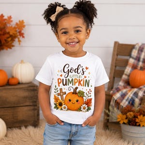 May include: A white short-sleeved t-shirt featuring a graphic print that reads God's LITTLE PUMPKIN in brown and orange text. The design includes a smiling orange pumpkin, a wooden cross, sunflowers, white daisies, and autumn leaves.