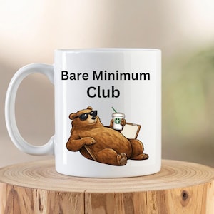 Puede incluir: Taza de cerámica blanca con el texto "Bare Minimum Club". La taza presenta un oso de dibujos animados con gafas de sol, sosteniendo una taza de café y descansando sobre una superficie de madera. El oso es marrón con gafas de sol negras.