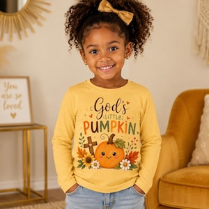 May include: A long-sleeve yellow shirt featuring a graphic of a smiling orange pumpkin, a wooden cross, sunflowers, and autumn leaves. The text "God's LITTLE PUMPKIN" is printed in brown and orange lettering above the pumpkin illustration.