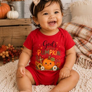 May include: A red short-sleeved baby bodysuit featuring a graphic of a smiling orange pumpkin, a wooden cross, sunflowers, and daisies. The text reads God's little PUMPKIN in black and orange lettering. A beige bow headband is worn on the head.