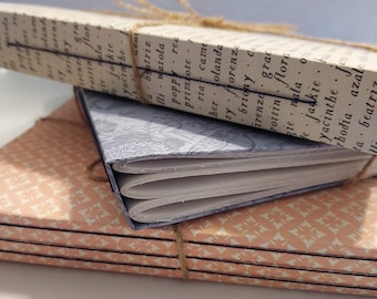 May include: A stack of four small, handmade journals with peach and white patterned covers. The top journal has a blue and white patterned cover and is tied with twine.