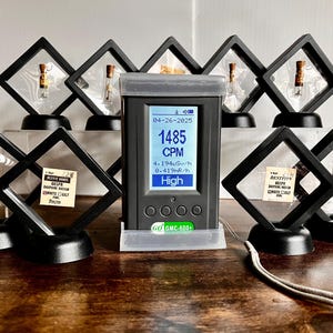 May include: A black digital radiation detector displays a reading of 1485 CPM, with a high radiation level indicator. It is surrounded by several black display cases, each containing a small vial. The text "GMC-600+" is visible on the detector.