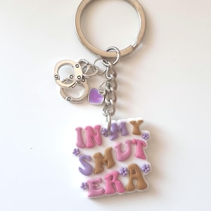 May include: A silver keychain with a ring, chain, and charms. Charms include handcuffs, a purple heart, and a rectangular charm with the words "IN MY SMUT ERA" in pink, brown, and purple letters, decorated with small flowers.