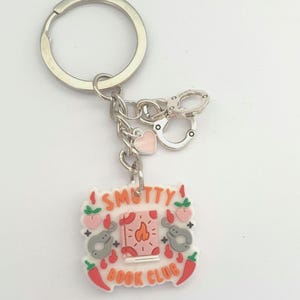 May include: Keychain with a silver ring, chain, and charms. The charms include a pink heart, handcuffs, and a rectangular charm with the text "SMUTTY BOOK CLUB". The rectangular charm has illustrations of a book, hearts, and chili peppers.